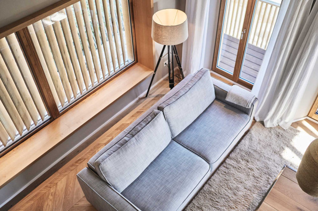 Boutique Apartment Bergfreuden - Zu sehen ist der Ausschnitt des Wohnzimmers von oben mit hellem, naturbelassenen Eiche Fischgrätparkett, mit hellgrauem Sofa, hellem flauschigem Teppich mit Couchtisch mit Tischplatte aus hellen Eichenholzdielen und ein Panoramafenster mit Holzlamellen (Photo 40)