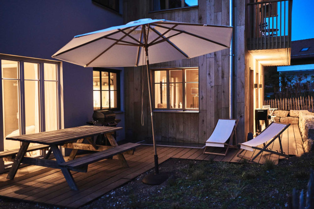 Boutique Apartment Flachsblüte - Ferienhaus mit Terrasse, auf der ein weißer Sonnenschirm und zwei weiße Deckchairs stehen, bei Dämmerung mit warmer Beleuchtung von innen (Photo 13)