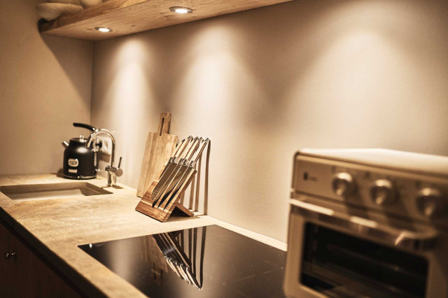 Das Flachshaus Ferienhaus - Küchenzeile mit Natursteinwaschbecken, Natursteinarbeitsplatte mit Messerblock aus Holz, schwarzem Wasserkocher, einem schwarz glänzenden Cerankochfeld, sowie einem kleinen beigen, nostalgischen Ofen (Photo 19)