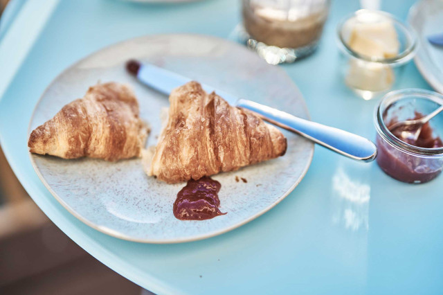 Das Flachshaus Ferienhaus - Handgefertigter hellblauer Keramikteller mit geteiltem Croissant, Marmelade und einem glänzendem, schlichten Edelstahlmesser auf hellblauer runder Tischplatte (Photo 21)
