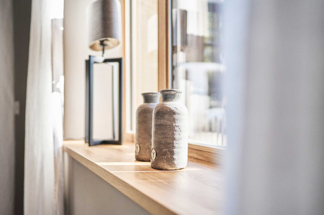 Boutique Apartment Flachsblüte - Fensterbank aus hellem Eichenholz mit zwei dekorativen, grauen Tonvasen. Im Hintergrund unscharf eine Tischleuchte mit schwarzem, filigranem Leuchtenfuß und grauem Leuchtenschirm (Photo 19)