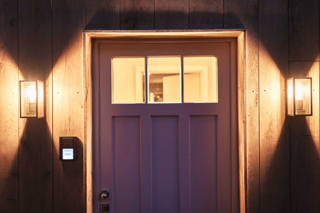 Das Flachshaus Ferienhaus - Zwei moderne schwarze Outdoor-Wandlaternen mit warmer Beleuchtung an Holfassade rechts und links neben Haustür mit Sprossenfenstern als Oberlicht, durch das warmes Licht scheint (Photo 38)