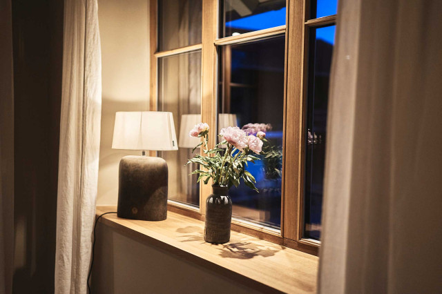 Das Flachshaus Ferienhaus - Dreiteiliges Sprossenfenster aus Holz mit Holzfensterbank, auf der eine warmleuchtende Tischleuchte mit weißem Leuchtenschirm und Leuchtenfuß aus Ton, sowie einer dunklen Tonvase mit rosafarbenen Blumen steht (Photo 39)