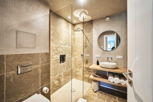 Das Flachshaus Ferienhaus - Badezimmer mit Naturstein aus Muschelkalk, Duschwand aus Glas, natürlichem Kalkspachtel, einem runden, beleuchteten Spiegel, sowie einem Waschtisch aus Eichebrettern und einem weißen Keramikwaschbecken (Photo 40)