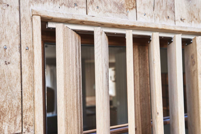 Boutique Apartment Flachsblüte - Holzlamellen aus hellem Eichenholz vor Holzsprossenfenster in heller Eichenholz-Fassade, im Hintergrund ist auf dem Bild schwach ein Raum erkennbar (Photo 25)