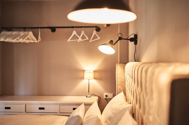 Boutique Apartment Bergfreuden - Bett mit hellem Chesterfieldbettrücken, weißer gefalteter Bettdecke und drei aufgestellte Kissen, zwei schwarze Industrialdesignwandleuchten mit warmer Beleuchtung, ein weißes Sideboard und eine schwarze Stahlrohrkleiderstange (Photo 30)