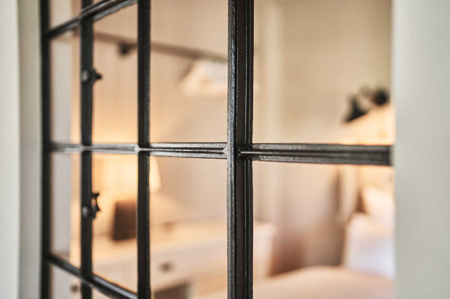 Boutique Apartment Flachsblüte - Im Bildfokus ist ein schwarzes Stahlsprossenfenster zu sehen, dahinter verschwommen ein hell bezogenes Bett mit hellbeigem Bettrücken und warme Beleuchtung (Photo 28)