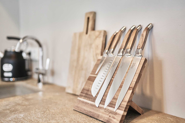 Boutique Apartment Flachsblüte - Im Bildfokus ist eine Küchenarbeitsplatte aus Naturstein in warmen, hellen Erdtönen, darauf ein Messerblock aus Eichenholz und Laguiole Küchenmesser, im Hintergrund etwas verschwommen ein schwarzer, nostalgischer Wasserkocher erkennbar (Photo 29)