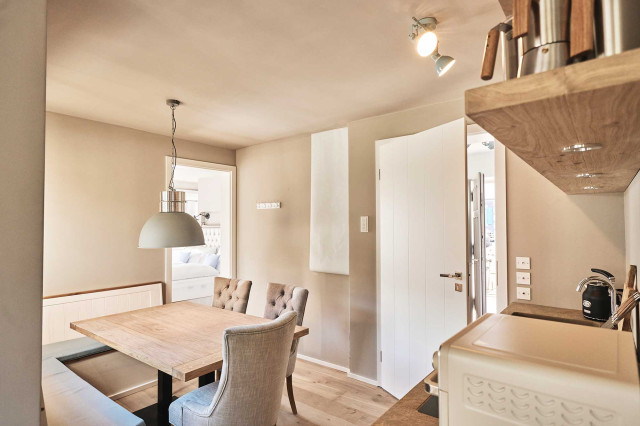Boutique Apartment Flachsblüte - Wohnküche mit gemütlicher heller Sitzecke, weißer Eckbank, einer Tischplatte aus hellem, massivem Eichenholz, großer hellgrauer Pendelleuchte in Industrial Design. Im Hintergrund ist ein helles Schlafzimmer mit frisch bezogenem Bett erkennbar (Photo 31)