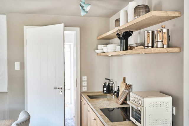 Boutique Apartment Flachsblüte - Blick seitlich auf Küchenzeile mit Natursteinarbeitsplatte, Natursteinspülbecken, Cerankochfeld, kleinem beigen Ofen, heller Eichenholz-Küchenfront und hellen Regalböden aus Eichenholz mit Keramikgeschirr, Vasen, Wasserkaraffen und Kochbesteck (Photo 35)