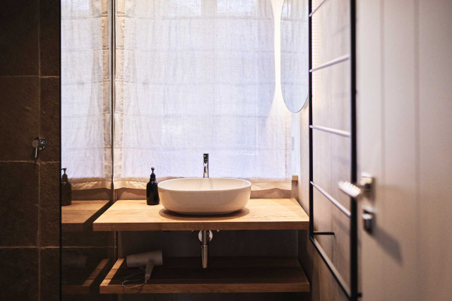 Boutique Apartment Bergfreuden - Badezimmer mit Eichenholzwaschtischbrettern mit ovalem weißen Keramikwaschbecken, schlichter Badarmatur, dahinter ein weißes heruntergelassenes Leinenrollo, links im Bild ist eine Natursteindusche zu sehen (Photo 29)