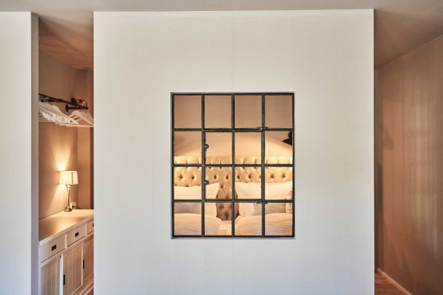 Boutique Apartment Flachsblüte - Hellgraue Trennwand mit schwarzem Stahlsprossenfenster, dahinter in warmem Licht ein Chesterfield-Design-Bett und eine Garderobenzeile mit schwarzer Kleiderstange aus Stahlrohr (Photo 6)