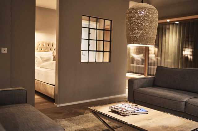 Boutique Apartment Bergfreuden - Offenes Wohn- Schlafzimmer mit warmer Beleuchtung und Raumteiler mit schwarzem Stahlfenster, dahinter ist das Boxspringbett mit hellem Chesterfielddesign Bettrücken und schwarze Industiraldesign Wandleuchten zu sehen (Photo 31)