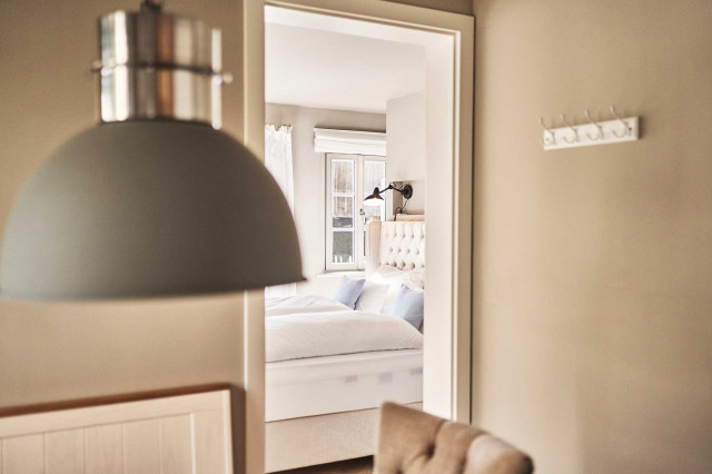 Boutique Apartment Flachsblüte - Im Vordergrund ist verschwommen eine graue Pendelleuchte in Industrial Design zu sehen. Im Hintergrund ein helles Bett in Chesterfield-Design mit weißer Bettwäsche. (Photo 40)