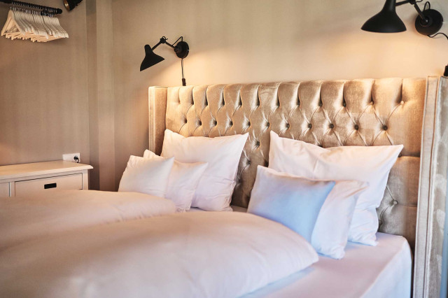 Boutique Apartment Bergfreuden - Schlafzimmer mit Boxspringbett, zwei gefalteten weißen Bettdecken, je drei stehende Kissen und beigem Chesterfielddesign Bettrücken, darüber zwei schwarze Wandleuchten im Industrialdesign, die eine warme Beleuchtung abgeben (Photo 32)