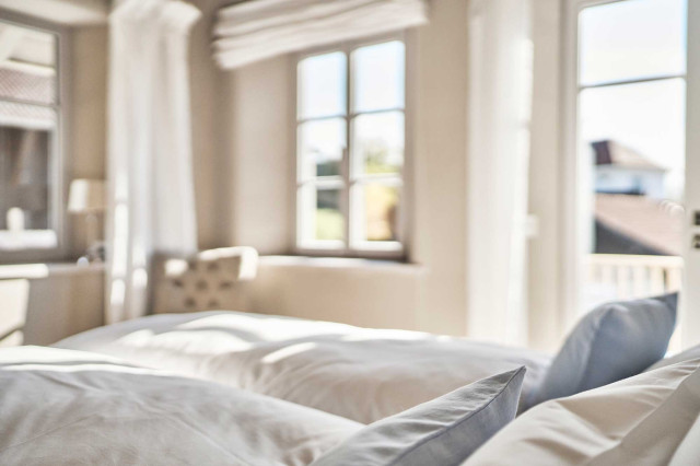 Boutique Apartment Bergfreuden - Schlafzimmer mit Boxspringbett mit zwei gefalteten weißen Decken, Fokus auf aufgestellten weißen Kissen u Dekokissen aus hellblauem Leinen, im Hintergrund verschwommen weiße Sprossenfenster, weiße Leinenvorhänge und ein Polstersessel im Chesterfielddesign (Photo 33)