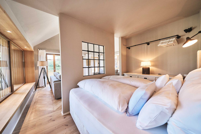 Boutique Apartment Bergfreuden - Offenes Schlaf- Wohnzimmer mit Trennwand, in der mittig ein schwarzes Stahlfenster zu sehen ist, Im Vordergrund ein Bett, links Panoramafenster mit Holzlamellen und Fischgrätparkett, im Hintergrund das Wohnzimmer (Photo 34)