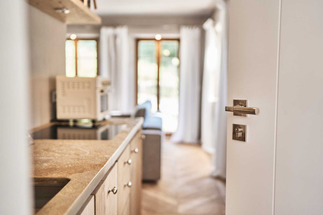 Boutique Apartment Flachsblüte - rechts im Bild eine weißlackierte Holztür mit Gropius Bauhaus-Klassiker Türgriff, links eine Küchenzeile mit Arbeitsplatte und Spülbecken aus Natursteins in warmen Erdtönen, im Hintergrund Eiche-Fischgrät-Parkett und ein Wohnzimmer verschwommen (Photo 43)