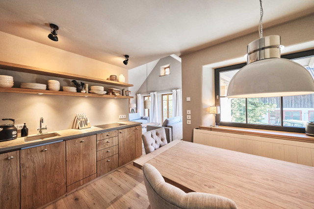 Boutique Apartment Bergfreuden - Im Vordergrund ist eine Wohnküche mit warmer Beleuchtung, Sitzecke, moderner heller Eichenholzfront, Natursteinarbeitsplatte, hellen Eichenholzregalböden mit handgefertigtem Keramikgeschirr zu sehen, im Hintergrund ein Wohnzimmer mit offenem Giebel (Photo 37)
