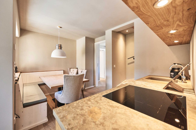Boutique Apartment Bergfreuden - Eine Wohnküche mit gepolsterter Sitzecke, gepolsterten Stühlen, einer Tischplatte aus massivem hellen Eichenholz und eine Natursteinarbeitsplatte in warmen Tönen, sowie ein schwarz glänzendes Ceranfeld (Photo 38)