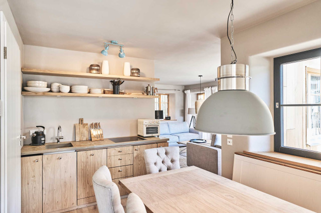 Boutique Apartment Flachsblüte - Wohnküche mit gemütlicher weißer Eckbank, massiver Eichenholz-Tischplatte, Küchenzeile mit heller, schlichter Eichenholz-Front, Natursteinarbeitsplatte und zwei Regalböden aus Eichenholz mit Keramikgeschirr und Vasen. Im Hintergrund ist ein Wohnzimmer. (Photo 46)