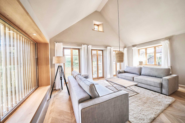 Boutique Apartment Bergfreuden - Wohnzimmer mit offenem Giebel, Panoramafenster mit Holzlamellen, hellem, naturbelassenen Eiche Fischgrätparkett, zwei hellgrauen Sofas, einem flauschigen Teppich, einem Couc