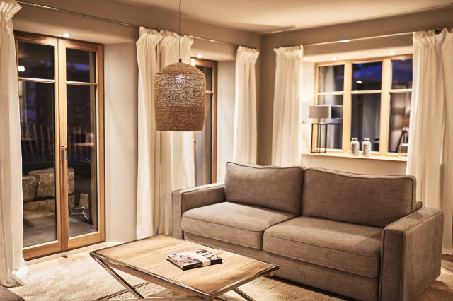 Boutique Apartment Flachsblüte - Wohnzimmer mit hellgrauem Sofa, flauschigem hell beigem Teppich und Holzsprossenfenstern mit Sturzleuchten, die eine warme Beleuchtung verströmen (Photo 9)