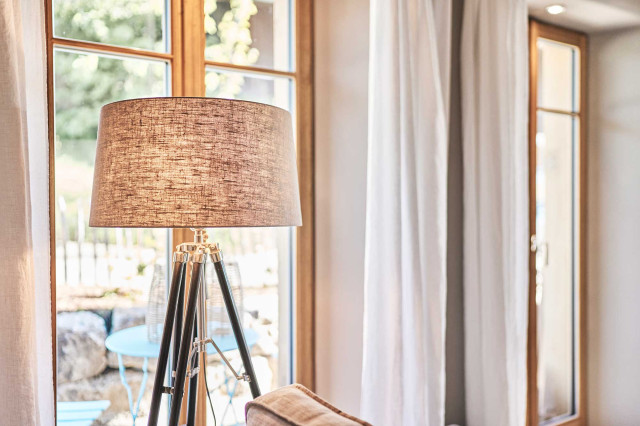 Boutique Apartment Flachsblüte - Tripodstehleuchte mit schlammfarbenem, durchschimmernden Leuchtenschirm und schwarzem Leuchtenfuß. Im Hintergrund verschwommen ein Holzfenster, durch das ein Baum mit grünen Blättern erkennbar ist (Photo 49)